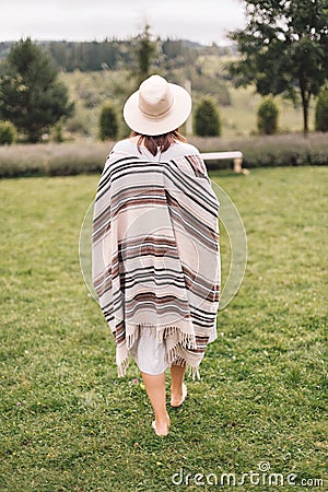 Stylish hipster girl in poncho, linen dress and hat walking at lavender field and relaxing in the morning. Bohemian woman enjoying Stock Photo