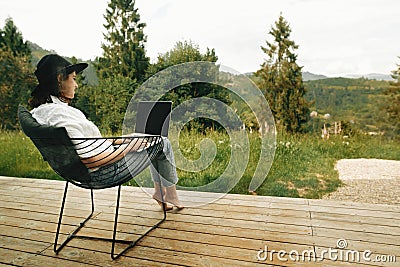 Stylish hipster girl sitting with laptop on terrace with view on woods. Young happy woman in hat using laptop, shopping or working Stock Photo