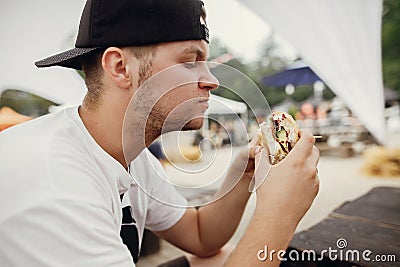 Stylish hipster man eating delicious vegan burger at street food festival. Hungry man biting burger with vegetables in summer Stock Photo