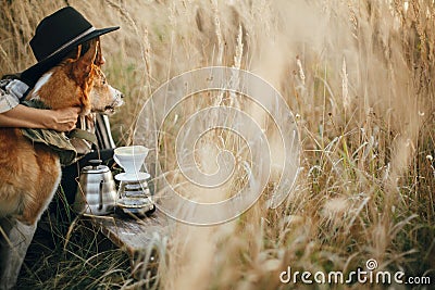 Stylish hipster traveler hugging her golden dog and brewing fresh coffee in rural countryside herbs in sunset light. Atmospheric Stock Photo