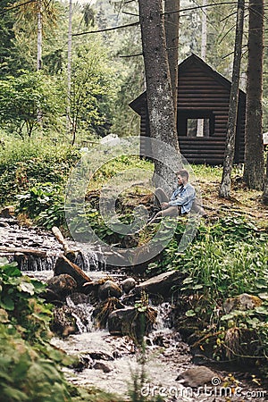 Stylish hipster traveler sitting at river in forest on background of wooden log in mountains. Exploring woods in summer. Copy Stock Photo