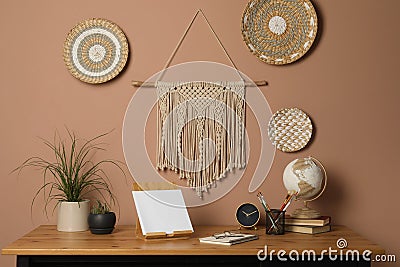 Stylish macrame and wicker wall decor hanging above wooden table with houseplants and stationery Stock Photo