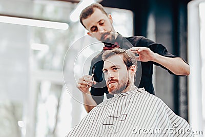 Stylish mustachioed barber dressed in a black shirt with a red bow tie scissors the hair of a young man in a barbershop Stock Photo