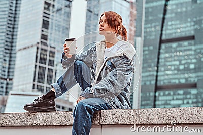 Stylish redhead hipster girl with tattoo on her face wearing denim coat holding takeaway coffee sitting in front of Stock Photo