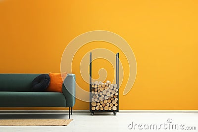 Stylish sofa with cushions, rug and firewood near orange wall indoors, space for text. Interior design Stock Photo