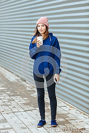 Stylish sporty brunette woman in trendy urban outwear posing with big white disposable cup straw cold rainy fall day against strip Stock Photo