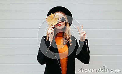 Stylish woman closes her eyes with yellow maple leaves blowing red lips sending sweet air kiss, female model in black coat Stock Photo