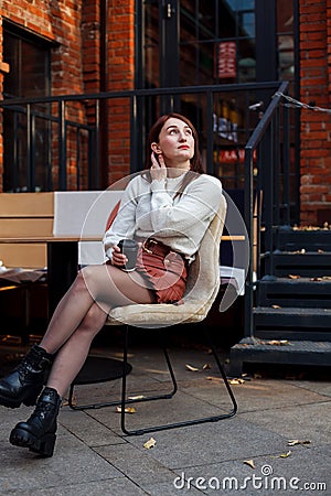 Stylish woman sitting on a cafe terrace with takeaway coffee and correcting her hairs while looking up Stock Photo