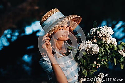 A stylish woman in a straw hat poses with a bouquet of lilacs, in a Sunny spring Park. A quiet portrait of a beautiful girl Stock Photo