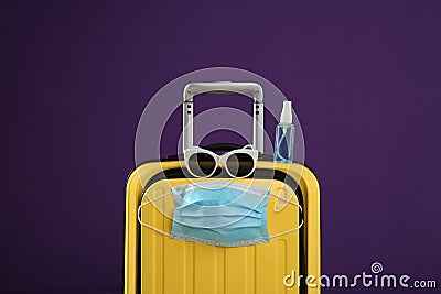 Stylish yellow suitcase with protective mask, sunglasses and antiseptic spray on purple background. Travelling during coronavirus Stock Photo