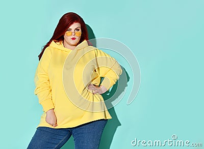 Stylish young fatty lady in glasses thoughtfully looks up winding strand of her red hair on finger. Three quarter length portrait Stock Photo