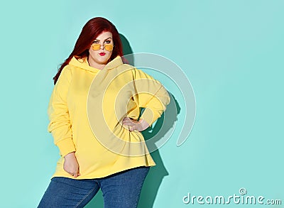Stylish young fatty lady in glasses thoughtfully looks up winding strand of her red hair on finger. Stock Photo