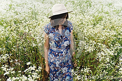 Stylish young woman in blue vintage dress and hat posing in summer meadow in many white wildflowers. Tranquil summer in Stock Photo