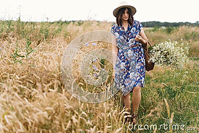 Stylish young woman in blue vintage dress and hat walking with white wildflowers in straw basket at yellow wheat field. Beautiful Stock Photo