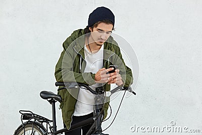 Stylish youngster in black hat and jacket with hood, has active lifestyle, covers long distances with his sport bicycle, has break Stock Photo