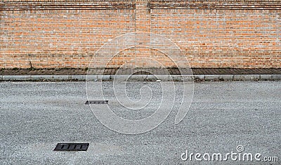 Suburban street background for copy space. Asphalt road with manholes in front of a sidewalk with weeds and a brickwall Stock Photo