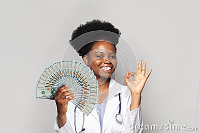 Successful african american woman doctor smiles and shows OK and holds fan of moneyagainst white background Stock Photo