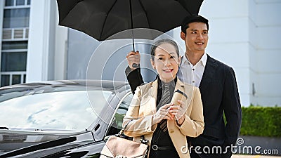 Successful Asian aged businesswoman with handsome male bodyguard under the umbrella Stock Photo