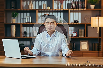 Successful businessman meditates sitting at a table in a classic office, Asian man in shirt relaxes, after working on laptop, man Stock Photo