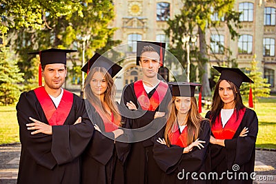 Successful confident five graduates in robes and hats standing in row while crossing hands Stock Photo