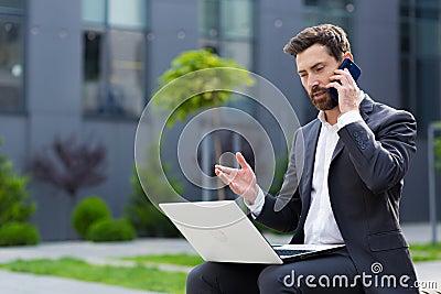 Successful male businessman reports good news by phone, working with laptop at lunchtime near office sitting on bench Stock Photo