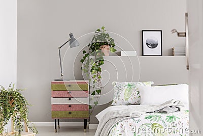 Suede covered pink and olive green nightstand with grey lamp in stylish bedroom with floral sheets and green plants in pots Stock Photo