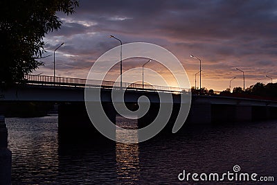 Summer evening with sunset sky, golden sunbeams, blue clouds on promenade, black silhouette bridge with lanterns lights over river Stock Photo