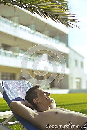 Summer holidays. Young man sunbathing. In the background a palm tree. Vacation concept. Man freelancer lies on a blue lounger. Stock Photo