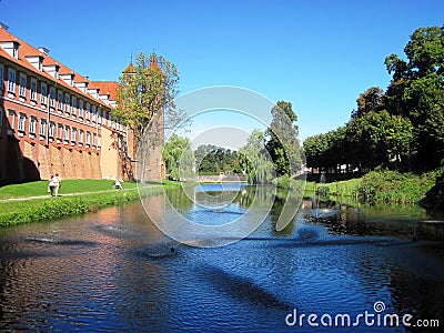 In summer, people walk along the bank of a moat with water around a medieval brick castle with towers. Stock Photo