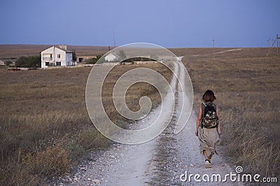 Summer steppe - a dark-haired girl with a backpack walks along the road that goes to the buildings of houses and beyond Stock Photo