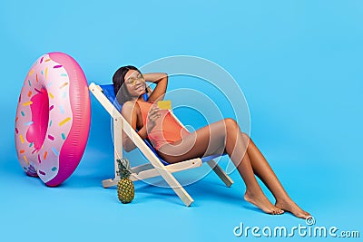 Summer vacation budget. Pretty black woman in suimsuit relaxing in lounge chair with credit card on blue background Stock Photo
