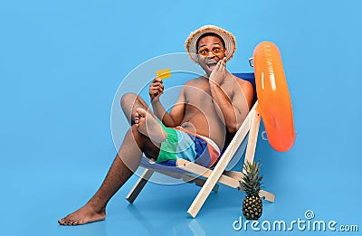 Summer vacation budget. Shocked black guy with credit card chilling in lounge chair, shouting OMG over blue background Stock Photo