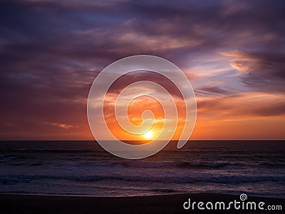 Sun on horizon at sunset with blue orange gradient sky and dark dramatic clouds over ocean Stock Photo
