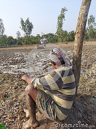 A farmer taking rest under a tree observing his son pulling tractor in the muddy field. Editorial Stock Photo