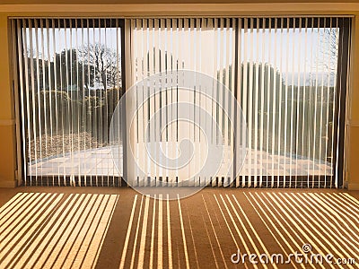 Sunlight shining through full length white vertical blinds in front of three glass sliding french doors leading to a patio. Stock Photo
