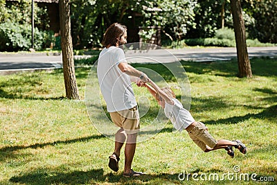 Sunny day young dark-haired father and his little son in white t shirts having fun and playing outdoor. Stock Photo