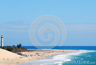 A sunny sand beach being hit gently by foamy wave crests, with a lighthouse standing on a pouch of tropical trees Stock Photo