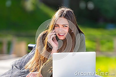 On a sunny and windy day young smiling woman lying on bench in park and working on laptop and talking on a smartphone Stock Photo