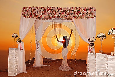 Sunset. bride silhouette. Wedding ceremony arch with flower arrangement and white curtain on cliff above sea, outdoor photo. Stock Photo