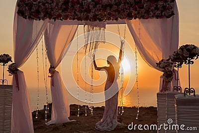 Sunset. bride silhouette. Wedding ceremony arch with flower arrangement and white curtain on cliff above sea, outdoor photo. Stock Photo