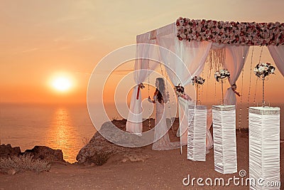 Sunset. bride silhouette. Wedding ceremony arch with flower arrangement and white curtain on cliff above sea, outdoor photo. Stock Photo