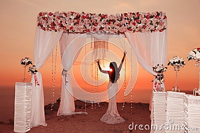 Sunset. Bride silhouette. Wedding ceremony arch with flower arrangement, white curtain and crystals chandelier on cliff above sea Stock Photo