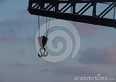 Sunset view of a metal double hook hanging with ropes from a tall crane in front of a cloudy blue pink sky Stock Photo