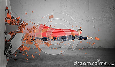 A superhero businessman in a cape flying right through the wall leaving rubble behind him. Stock Photo