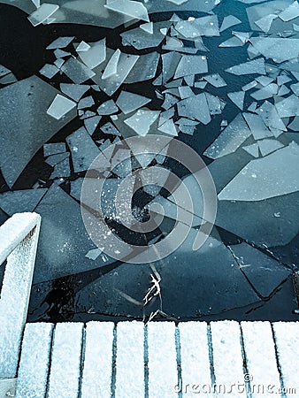 Surface of the lake with cracked ice. Black water with frozen parts. Wooden boards under snow. Stock Photo