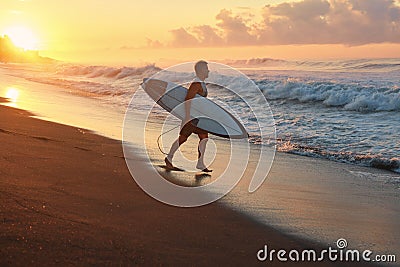 Surfer. Surfing Man With White Surfboard Going To Surf On Ocean Waves. Sandy Beach At Beautiful Sunrise In Bali. Stock Photo