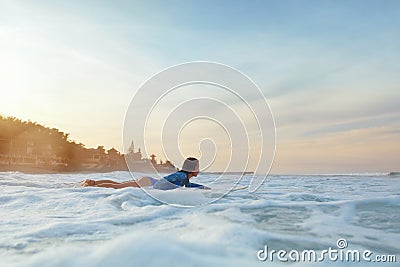 Surfing Girl. Surfer Woman On Surfboard In Ocean. Brunette In Blue Wetsuit Swimming In Sea At Sunset. Stock Photo