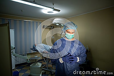 Surgeon in blue gloved thinking about surgery. The patient lies on the couch in the background. Preparation for surgery Stock Photo