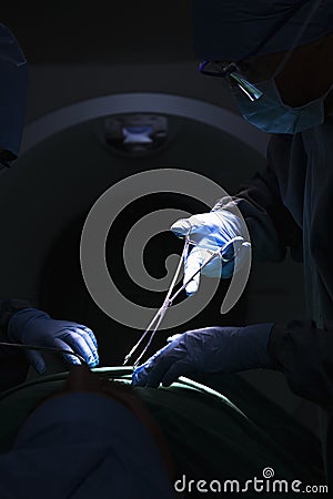 Surgeon looking down, working, and holding surgical equipment with patient lying on the operating table, dark Stock Photo