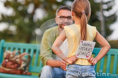 Surprise! Rear view of cute little girl hiding handmade postcard Happy Fathers Day behind her back while greeting her Stock Photo